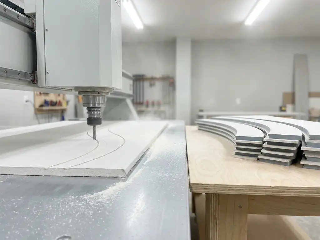 CNC-Fräsmaschine schneidet geschwungene Profile in eine weiße Gipskartonplatte, feiner Staub auf hellem Werkstatttisch.