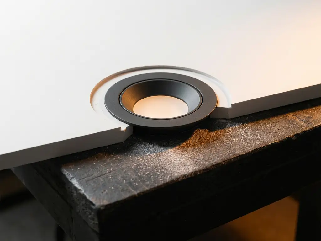 LED-Einbaustrahler in kreisrunder Aussparung einer weißen Gipskartonplatte, weißer Staub auf dunkler Werkbank, warmes Amberlicht.